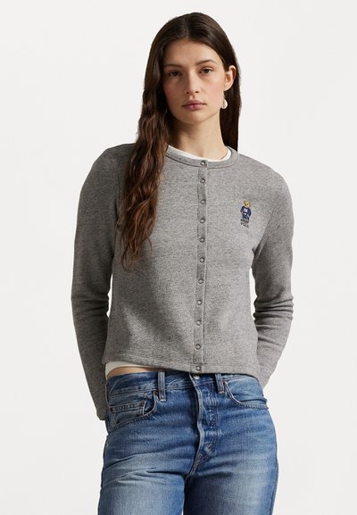 Cardigan grigio con scollatura rotonda, chiusura frontale con bottoni e ricamo di un orso. Maniche lunghe, texture morbida, abbinato a jeans di denim.