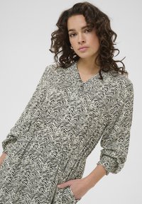 Rochie cu mânecă lungă, cu un model ondulat în negru și crem, având un guler smockat și mansete elastice, cu o talie plisată și buzunare.