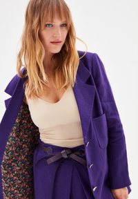 Blazer di lino viola con un rivestimento a motivo floreale, abbinato a un top beige a coste e pantaloni a vita alta con una cintura in pelle.