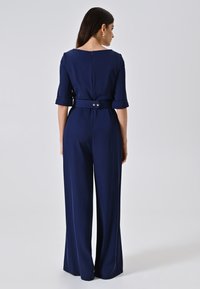 Navyblauwe jumpsuit met korte mouwen, een taillering en riemdetail. Glad materiaal, wijde pijpen, sluiting met rits aan de achterkant en gouden knopen.