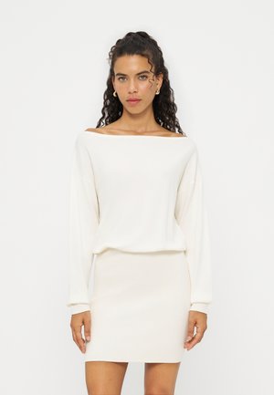 Femme aux cheveux bouclés portant un top blanc à manches longues épaules dénudées et une jupe blanche côtelée assortie, debout devant un fond uni.