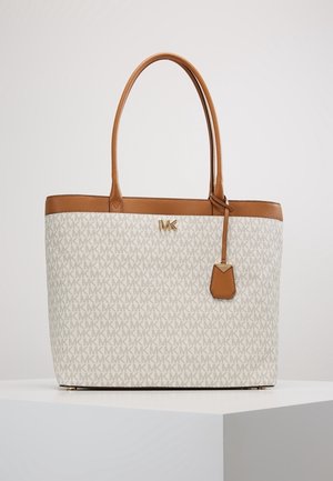 Beige en bruine Michael Kors shopper met herhaald MK-patroon, leren handvatten en een klein hangend leren label aan een wit voetstuk.