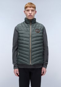 Dunkelgrüne Steppweste mit Zickzacksteppung, durchgehendem Reißverschluss, Stehkragen und Logopatch; über einem grauen Pullover getragen.