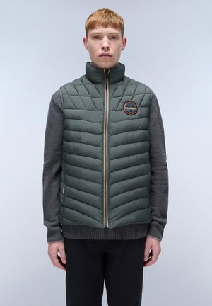 Polstret vest i mørkegrøn med chevron-quilting, hel lynlås, opretstående krave og logo-patch; båret over en grå sweater.