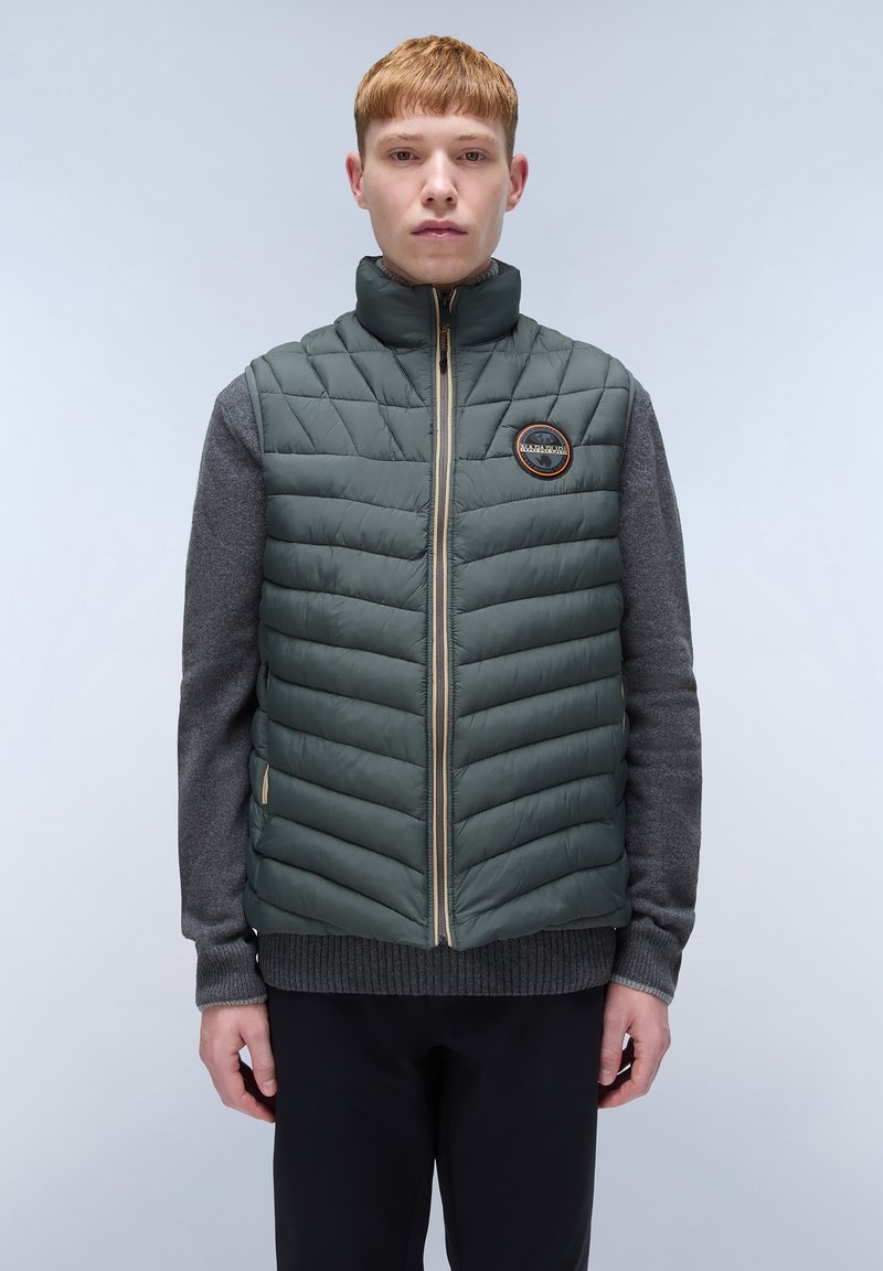 Dunkelgrüne Steppweste mit Zickzacksteppung, durchgehendem Reißverschluss, Stehkragen und Logopatch; über einem grauen Pullover getragen.