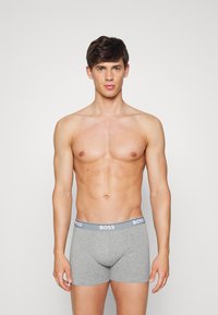 Graue Baumwoll-Boxershorts mit einer enganliegenden Passform, ausgestattet mit einem weißen Bund, der mit "BOSS" beschriftet ist, sowie einer glatten Textur. Das Modell steht aufrecht.