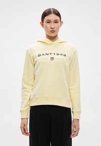 Žltá mikina s kapucňou z jemného materiálu. Na prednej strane je logo "GANT 1949" s doplnkovým textom a erbovým emblémom. Dlhé rukávy a kapucňa so šnúrkou.