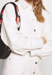 Giacca bianca con bottone a pressione e vestibilità rilassata, caratterizzata da bottoni metallici, tasche oversize e una tracolla nera con accenti in rosso e bianco del logo.