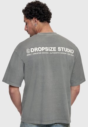 Mand med krøllet hår og øreringe bærer en grå dropsize studie t-shirt med hvid tekst og blå jeans, vendt væk mod en ensfarvet baggrund.