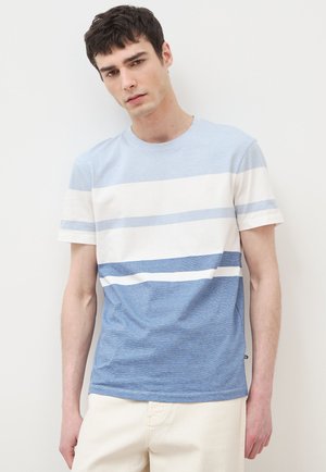 Junger Mann mit kurzem dunklem Haar trägt ein blau-weiß horizontal gestreiftes T-Shirt und helle Hosen vor einem schlichten Hintergrund.