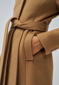 Cappotto di lana marrone con cintura e tasca; presenta una texture liscia, una silhouette dritta e bottoni decorativi sul polsino.