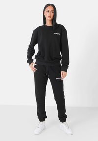 Completo felpa nera composto da felpa e pantaloni, realizzato in tessuto morbido. Presenta polsini a costine, vita con cordoncino e dettagli in logo bianco.