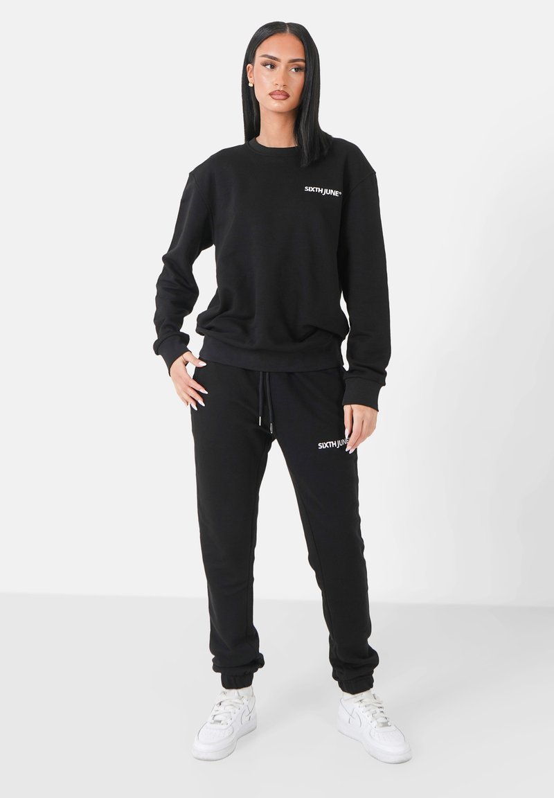 Completo felpa nera composto da felpa e pantaloni, realizzato in tessuto morbido. Presenta polsini a costine, vita con cordoncino e dettagli in logo bianco.