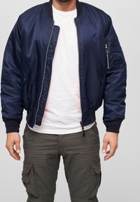 Námořnická modrá bomber bunda s lesklým povrchem, žebrovanými manžetami a kapsou na zip na rukávu, nošena přes bílý tričko.