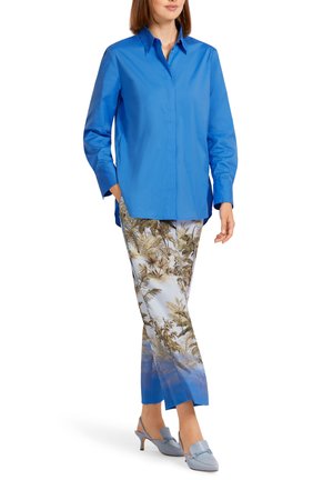 Femme portant une chemise boutonnée bleu cobalt, un pantalon court à imprimé tropical avec des ourlets dégradés bleus, et des chaussures à talons bleu clair à bride arrière.
