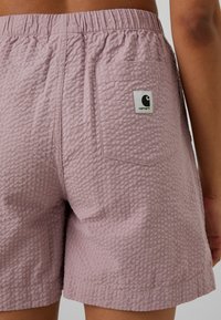 Shorts texturés violet clair avec une ceinture élastique et un patch logo Carhartt blanc carré sur la poche arrière, portés par une personne.