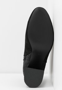 Semelle de chaussure noire avec talon texturé et avant lisse, reposant contre une surface d’angle blanche.