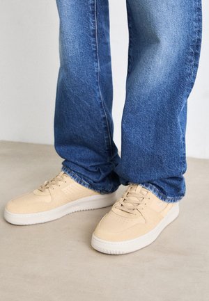 Blauwe spijkerbroek gedragen met beige veter sneakers op een lichte vloer, gefotografeerd vanaf halverwege de kuit naar beneden.