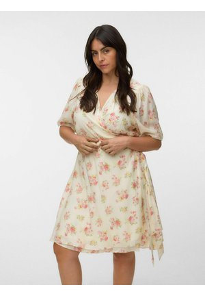 Donna con i capelli lunghi e scuri che indossa un vestito a portafoglio color crema chiaro con stampa floreale rosa e verde, in piedi davanti a uno sfondo bianco neutro.