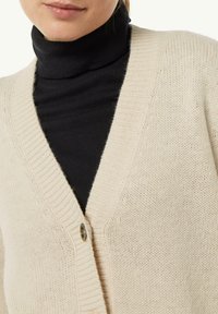 Beżowy sweter z dzianiny z dekoltem w kształcie litery V, z pojedynczym zapięciem na guzik i ribbowanym wykończeniem krawędzi. Nosi się go na czarnym golfie.