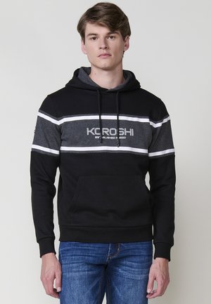 Joven con una sudadera con capucha negra y gris y un logo, de pie contra un fondo claro. Tiene el cabello corto y una expresión neutra.