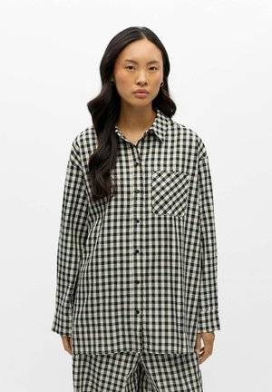 Jeune femme aux longs cheveux foncés portant une chemise à carreaux noire et blanche boutonnée et un pantalon assorti, debout devant un fond blanc.