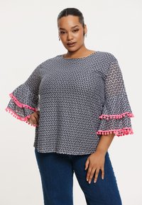 Schwarz-weiß gemusterte Bluse mit ausgestellt Ärmeln, verziert mit rosa Pompon-Bordüre entlang der Kanten. Leichtes Material, lockere Passform.