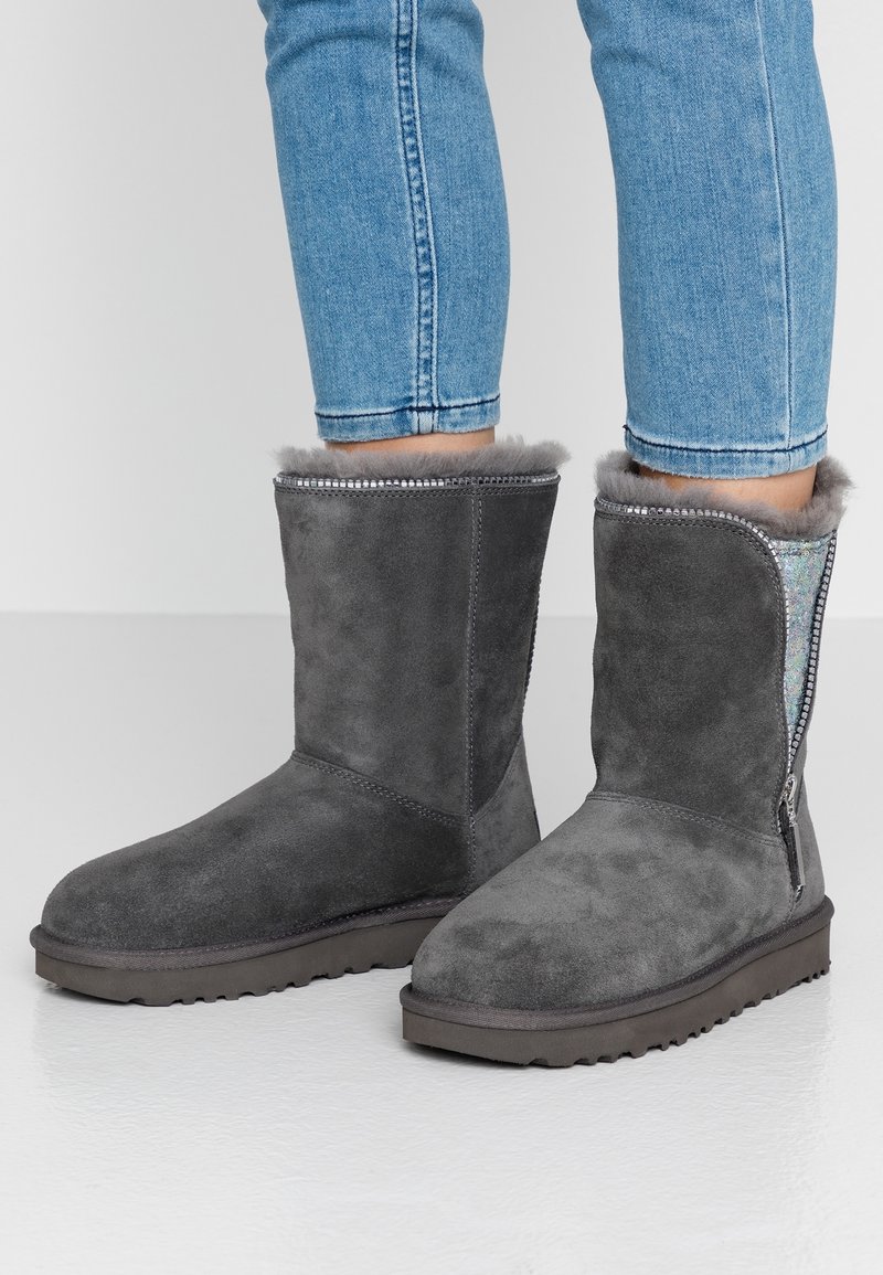 Bottes mi-mollet en daim gris avec doublure en fourrure et détail de fermeture éclair, portées avec un jean bleu clair sur une surface claire unie.