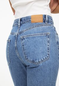 Acercamiento de una persona que lleva puesto un pantalón de mezclilla azul con una etiqueta de cuero de la marca y una camiseta blanca, mostrado desde la cintura trasera hasta la mitad del muslo.
