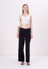 Bílý vykrojený top s volánovými ramínky, spárovaný s černými širokými kalhotami s vysokým pasem. Modelka drží malou bílou tašku a nosí černé sandály.