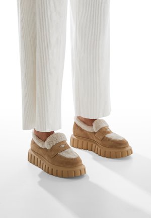 Zapatos de ante marrón con un suave forro de crema, que presentan un acento de corte y suelas de goma gruesas y estriadas. Combinados con pantalones blancos de canalé.