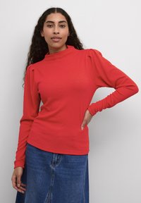 Maglietta rossa a maniche lunghe con colletto alto, tessuto texturizzato e maniche arricciate, abbinata a una gonna di jeans blu. Design semplice e aderente.