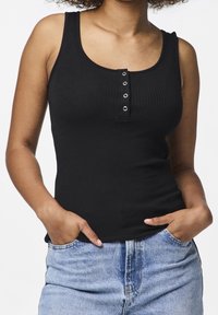 Svart ribbat linne med rundad halsringning och klädd knäppning, kombinerat med urtvättade blå jeans, händerna i fickorna, fokus på designen av linnet.