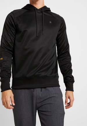 Uomo che indossa una felpa nera con cappuccio e coulisse a maniche lunghe e pantaloni grigio scuro, in piedi davanti a uno sfondo chiaro e uniforme.