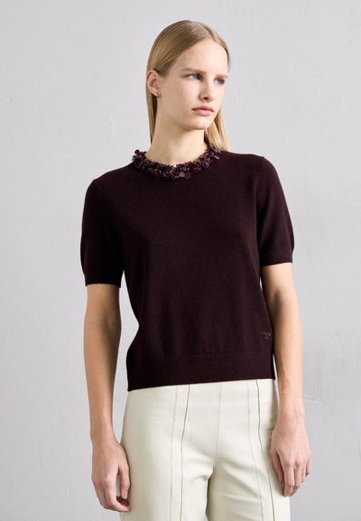Kortærmet, burgundy striktrøje med en tekstureret blomsterperlekant. Matchet med cremede, tætsiddende bukser med søm detaljer.