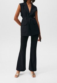 Schwarzer ärmelloser Blazer mit tiefem V-Ausschnitt, zwei Fronttaschen, tailliertem Bund, kombiniert mit schwarzen ausgestellten Hosen und metallischen Riemchensandalen.
