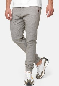 Pantalon de jogging gris clair en tissu texturé, avec poignets élastiques, poches avant et coutures contrastées. Porté avec des baskets blanches et grises.