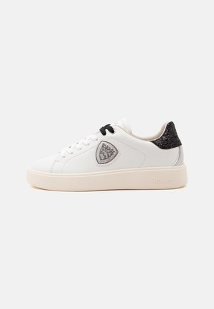 Sneaker bianca basse con toppa posteriore nera, lacci neri e bianchi, logo a scudo sul lato e suola spessa color panna.
