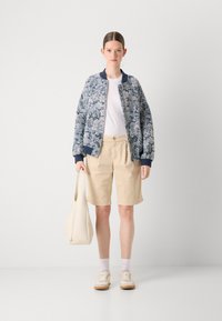 Veste bomber avec un motif floral en bleu et blanc, associée à un short beige. L'accessoire comprend un sac beige clair, porté avec des baskets blanches.
