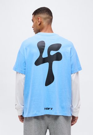 Homme vu de dos portant une chemise bleu clair avec un grand logo noir "4f" et un petit texte "10f1", superposée à une chemise blanche à manches longues.