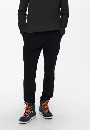 Black trousers made of soft fabric, relaxed fit. Paired with navy hiking boots featuring orange laces and silver eyelets.