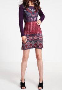 Langekruid jurk met een getailleerd ontwerp, gekenmerkt door kleurrijke geometrische patronen en gestippelde texturen, in tinten paars, rood en zwart.