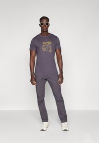 Γκρίζα μπλούζα τύπου t-shirt με κίτρινο γραφικό "Mountain Living", συνδυασμένη με αντίστοιχους γκρι παντελόνες και λευκά sneakers. Το μοντέλο φορά γυαλιά ηλίου.