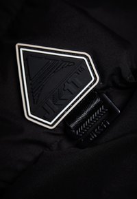 Tissu noir avec un patch triangulaire en caoutchouc noir texturé, contourné en blanc, portant l'inscription "JK1T" en gras. Comprend une sangle noire avec des détails.