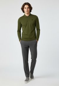 Junger Mann steht mit den Händen in den Taschen, trägt ein olivgrünes Langarmpolo, graue Hose und graue Sneakers vor einem weißen Hintergrund.