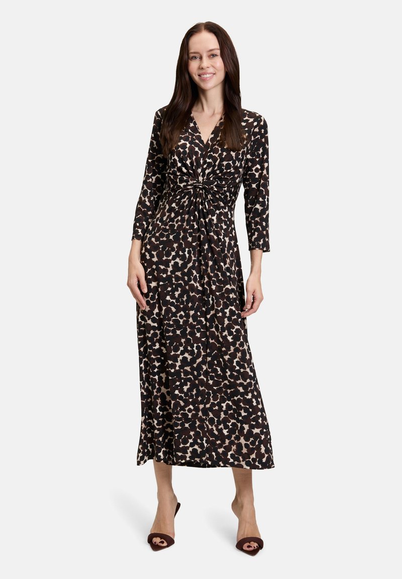 Femme souriante portant une robe portefeuille à manches longues, noire et beige à motif, avec un détail noué, se tenant sur des talons ouverts sombres sur fond blanc.