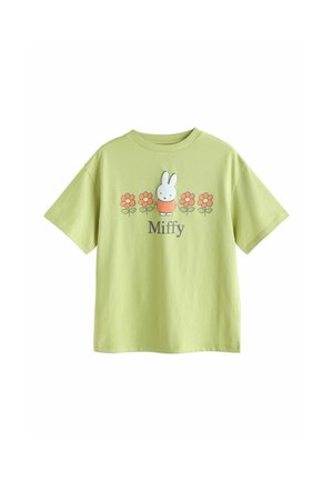 Fată cu păr împletit poartă tricou verde Miffy cu flori portocalii, asortat cu blugi largi bleu, stând în fața unui perete simplu bej.