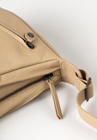 Borsa a tracolla in pelle beige con chiusura a zip, cuciture visibili e tracolla piatta. Presenta un bottone e una texture liscia.