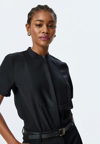 Blusa negra con un diseño estructurado, que presenta un panel de satén, mangas cortas, un cuello mao y un detalle de cierre en la parte delantera.