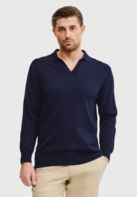 Granatowy sweter typu pullover z długim rękawem i dekoltem w kształcie litery V, o miękkiej fakturze, charakteryzujący się minimalistycznym wzornictwem oraz ściągaczami przy mankietach i u dołu.